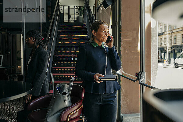 Reife Geschäftsfrau  die einen Terminkalender hält  während sie mit einem weiblichen Kollegen in einer Hotellounge über ein Smartphone spricht