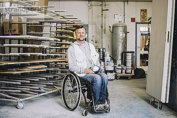 Lächelnder Maler in voller Länge  der einen Gehörschutz trägt  während er auf einem Rollstuhl in einer Werkstatt sitzt