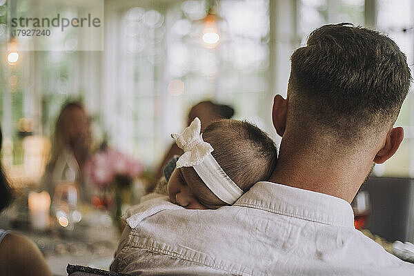 Tochter mit Stirnband schläft auf der Schulter des Vaters bei einer Dinnerparty