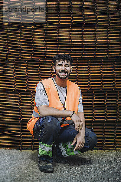 Ganzes Porträt eines glücklichen jungen Bauarbeiters  der auf der Baustelle hockt
