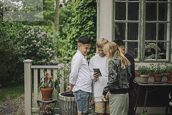 Lächelnde männliche und weibliche Freunde teilen sich ein Mobiltelefon  während sie in der Nähe eines Hauses stehen