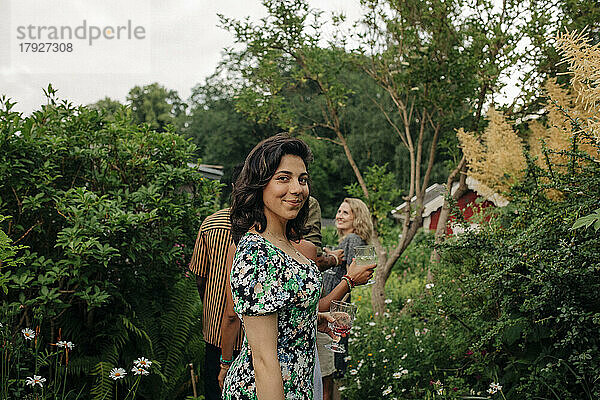 Seitenansicht einer lächelnden Frau mit Getränk  die bei Pflanzen im Garten steht