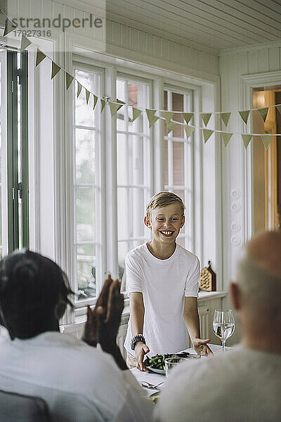 Lächelnder Junge serviert Essen während einer Dinnerparty zu Hause