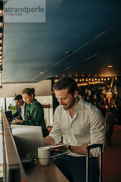 Lächelnder junger Geschäftsmann schreibt in Tagebuch  während er mit Laptop im Hotel sitzt