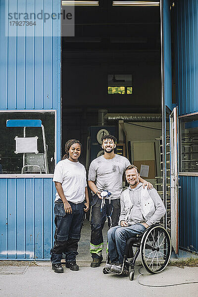 Lächelnder Maler und lächelnde Malerin in voller Länge mit einem Kollegen im Rollstuhl vor der Werkstatt
