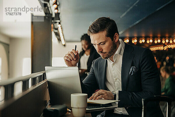 Junger Geschäftsmann liest Tagebuch  während er mit Laptop im Hotel sitzt