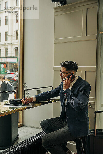 Geschäftsmann  der am Tisch in der Hotellounge sitzt und schreibt  während er mit seinem Smartphone telefoniert