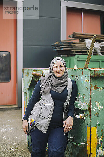 Lächelnde Bauarbeiterin mit Kopftuch lehnt sich an einen Metallcontainer