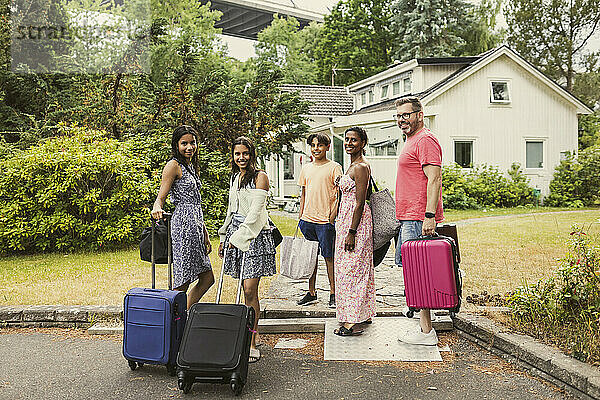 Porträt einer glücklichen gemischtrassigen Familie  die mit Gepäck vor einem Haus steht