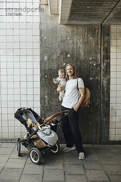 Lächelnder Mann mit Hand in der Tasche  der seine Tochter im Arm hält  während er mit dem Kinderwagen vor einer Mauer steht