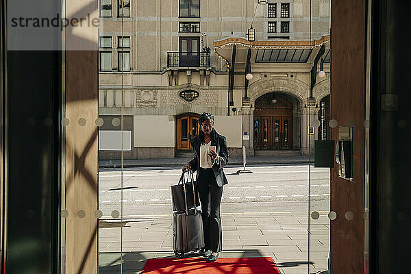 Junge Geschäftsfrau  die ein Smartphone benutzt  während sie mit einem Koffer vor einem Hotel in der Sonne steht