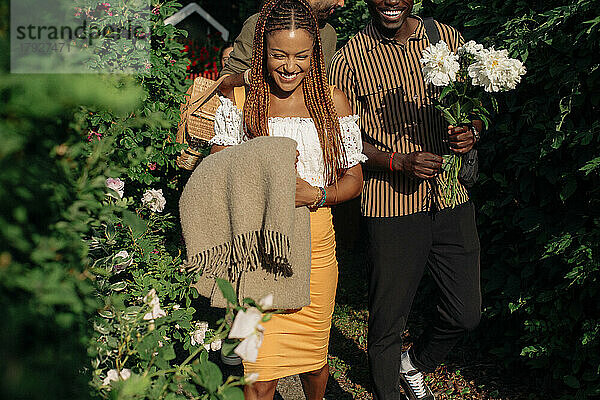 Lächelnde Frau  die eine Decke hält  während sie mit einem männlichen Freund bei Pflanzen an einem sonnigen Tag spazieren geht