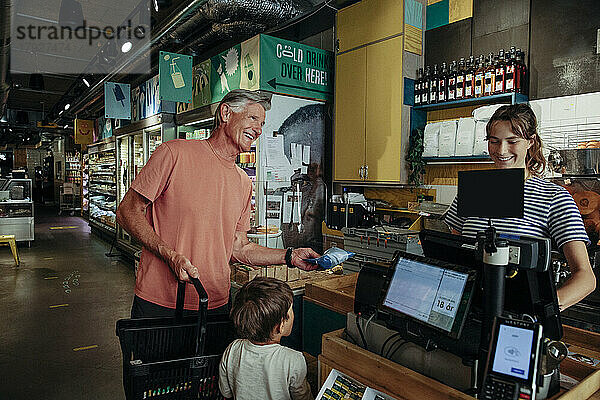 Glücklicher älterer Mann im Gespräch mit der Kassiererin  während er mit seinem Enkel an der Kasse im Supermarkt steht