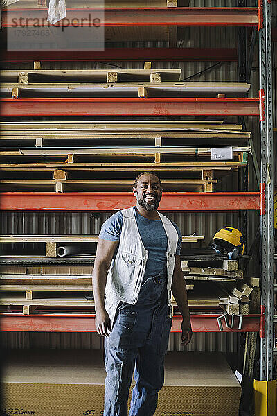 Porträt eines lächelnden Zimmermanns  der in einem Lagerhaus vor einem Palettenstapel steht