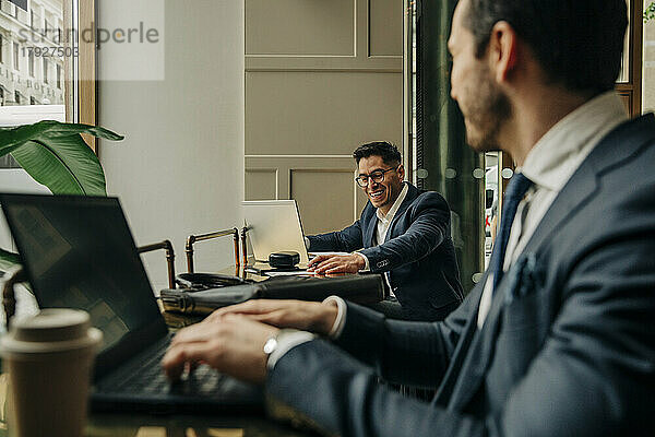 Glücklicher Geschäftsmann im Gespräch mit einem männlichen Kollegen  der einen Laptop benutzt  während er in einer Hotel-Lounge sitzt