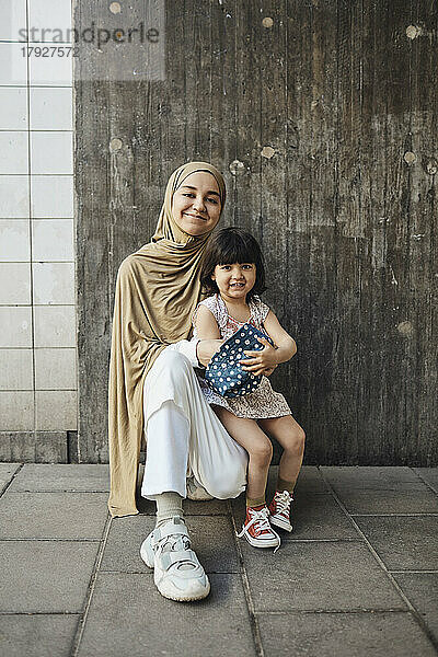 Lächelnde Frau mit Hidschab  die mit ihrer Tochter vor einer Mauer kniet