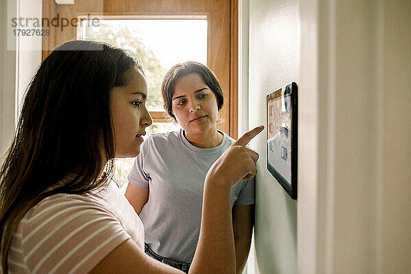 Mutter bringt ihrer Tochter bei  wie man die Hausautomatisierung auf einem digitalen Tablet nutzt  das zu Hause an der Wand montiert ist