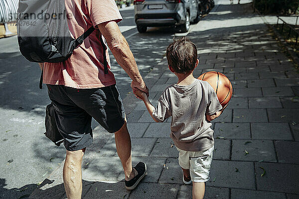 Rückansicht eines Mannes  der die Hand seines Enkels beim Gehen auf dem Bürgersteig hält