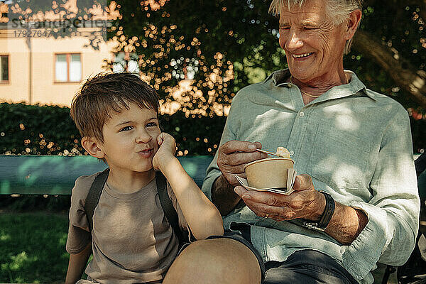Junge  der sich auf den Ellbogen stützt  während er mit seinem Großvater auf einer Bank sitzt und einen Eisbecher hält