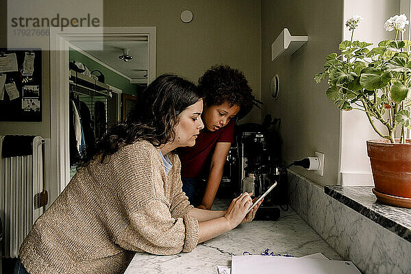 Mutter  die ihr Smartphone mit ihrem Sohn teilt  während sie sich zu Hause auf die Küchentheke stützt