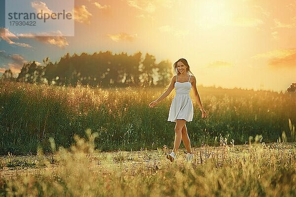 Glückliche junge Frau  die bei Sonnenuntergang auf einer Wiese entlang der Landstraße spaziert