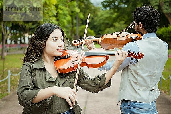 Zwei junge Geiger stehen und spielen Geige in einem Park. Porträt eines Mannes und einer Frau  die zusammen im Park Geige spielen. Violinist Mann und Frau Rücken an Rücken spielen Geige in einem Park im Freien