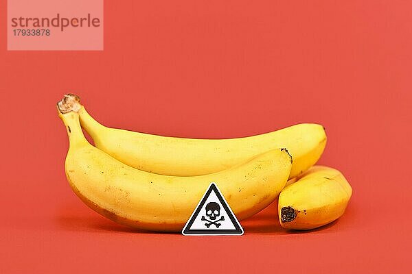 Konzept für ungesunde oder giftige Stoffe in Lebensmitteln wie Pestizidrückstände mit Totenkopf Warnschild vor Bananenfrüchten auf rotem Hintergrund