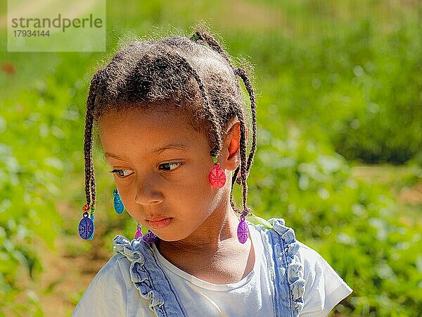 Afrikanisches kleines Mädchen im Gemüsegarten auf dem Bauernhof  das sich um die Pflanzen kümmert und im Sommer eine Jeans-Salopette trägt