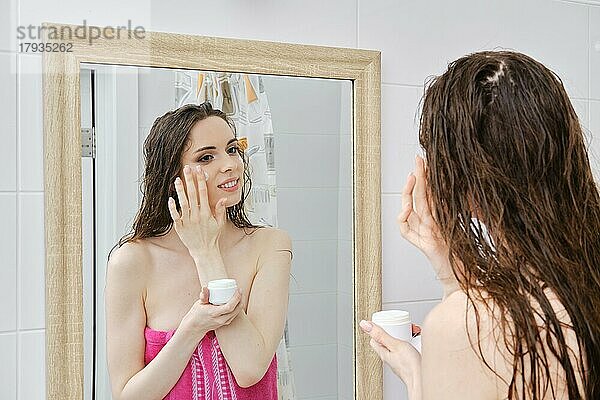 Junge lächelnde attraktive Frau in Handtuch eingewickelt Anwendung Creme auf Gesicht Haut  charmante weibliche stehend vor dem Spiegel im Bad und genießen tägliche Hautpflege Schönheitsprogramm. Schönheit Konzept