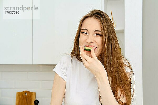 Lustige junge fröhliche Frau in weißem T-Shirt beißt Scheibe Gurke  schielt und lächelt. Glückliche Frau hat Spaß beim Kochen in der modernen weißen Küche zu Hause