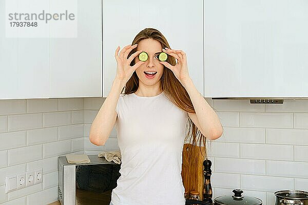 Lustiges Foto einer jungen  fröhlichen Frau in einem weißen T-Shirt  die ihre Augen mit Gurkenscheiben bedeckt und in die Kamera lächelt. Sie hat Spaß beim Kochen in einer modernen  weißen Küche zu Hause