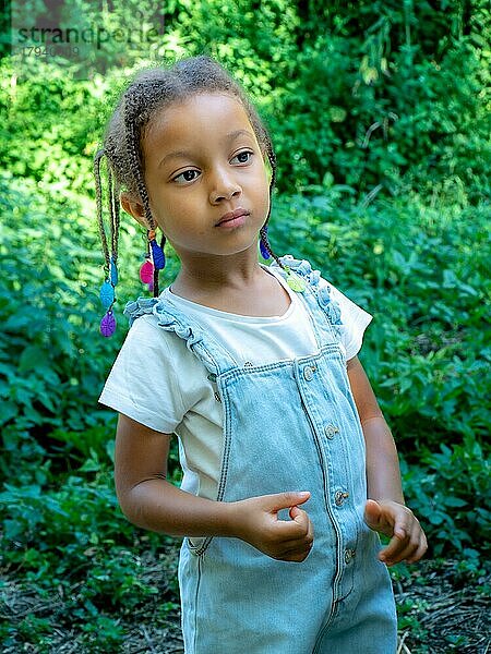 Afrikanisches kleines Mädchen im Gemüsegarten  das sich um die Pflanzen kümmert  mit Zöpfen und Jeans-Salopette im Sommer