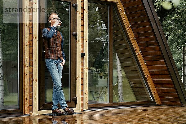 Selbstbewusster Mann mittleren Alters steht auf der Terrasse eines Holzchalets und lehnt sich gegen eine Glastür