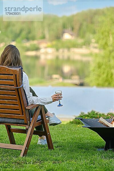 Junge Frau mit einem Glas Sangria in der Hand entspannt sich in der Natur neben einem Grill sitzend  Rückansicht