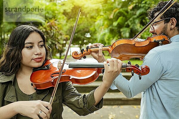 Geiger Mann und Frau Rücken an Rücken spielen Geige in einem Park im Freien. Zwei junge Geiger stehen und spielen Geige in einem Park. Porträt von Mann und Frau zusammen spielen Geige im Park