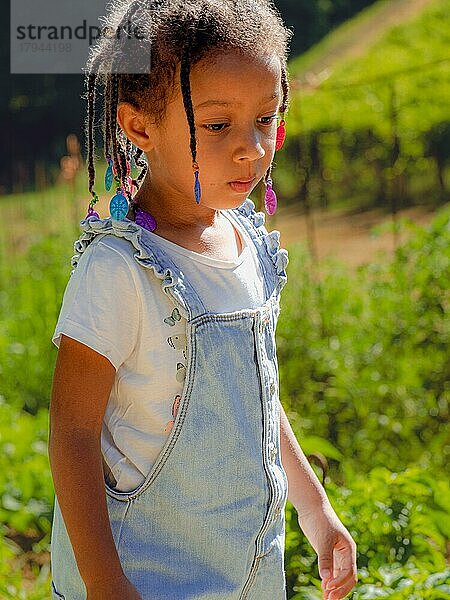 Porträt eines kleinen afrikanischen Mädchens  das im Sommer im Gemüsegarten des Bauernhofs spazieren geht und sich um die Pflanzen kümmert und dabei eine Jeans-Salopette trägt