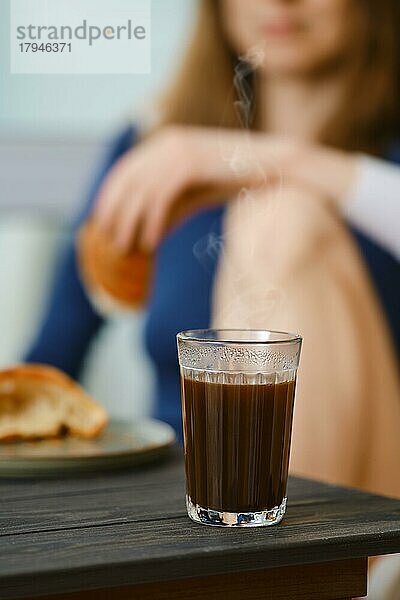 Kaffeeglas mit unkenntlicher  verschwommener Frau  die in der Küche sitzt und ein Croissant in der Hand hält  Fokus auf Glas mit Kaffee