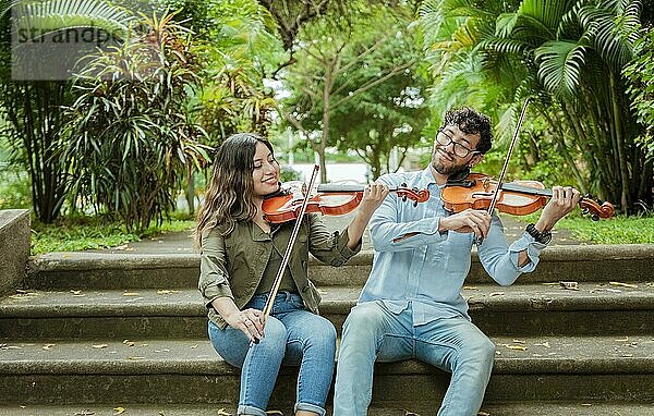 Porträt von zwei jungen Violinisten  die auf einer Treppe sitzen und Geige spielen. Porträt eines lächelnden Geigers  der auf einer Treppe sitzt und Geige spielt. Mann und Frau sitzen auf einer Treppe und spielen Geige