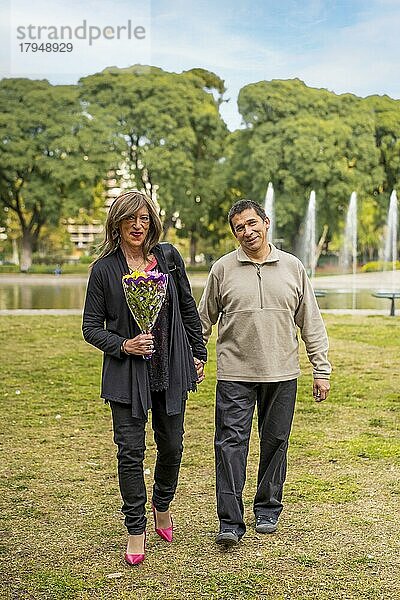 Reife Transgender Frau  die Hand in Hand mit ihrem Freund in einem Park spazieren geht und sich freut  weil er ihr Blumen geschenkt hat