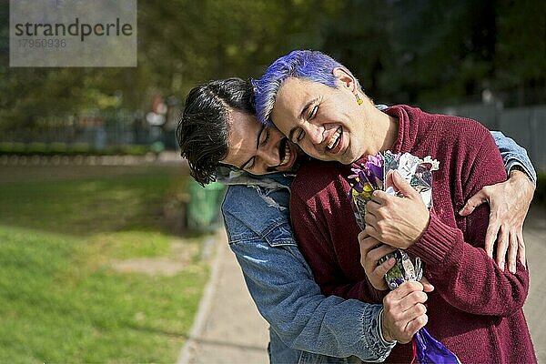 Ein schwules Latino Paar umarmt sich in einem Park  Einer überrascht den anderen mit einem Blumenstrauß als Geschenk