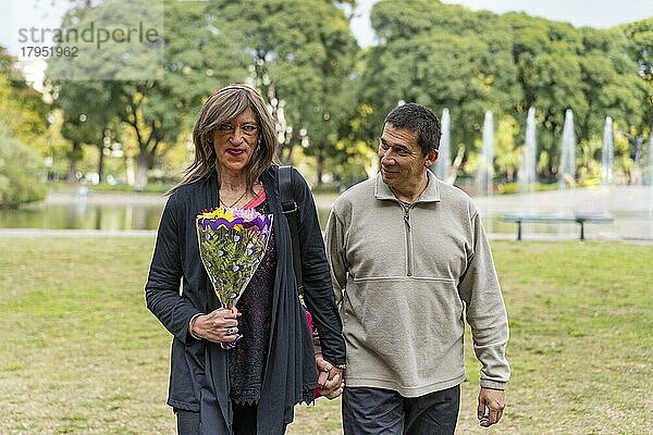 Reife Transgender Frau  die Hand in Hand mit ihrem Freund in einem Park spazieren geht und sich freut  weil er ihr Blumen geschenkt hat