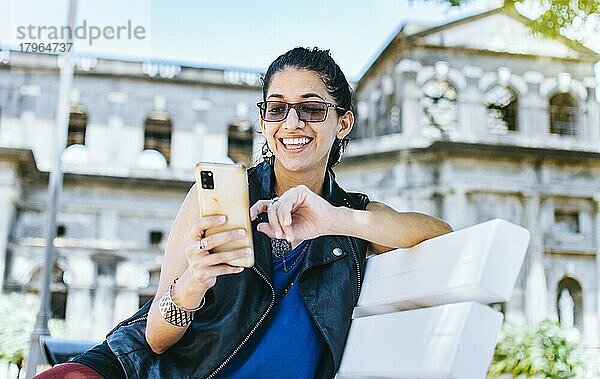 Ein attraktives Mädchen sitzt auf dem Handy lächelnd  Girl sitting on a bench texting on her cell phone  Urban style girl sitting on a bench with her cell phone
