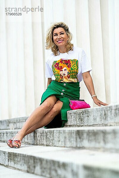 Hübsche Frau im besten Alter mit langen lockigen blonden Haaren sitzt auf einer Treppe und genießt die Wärme des Frühlings. Das weiße Shirt mit buntem Aufdruck und der kurze grüne Rock passt zur Jahreszeit