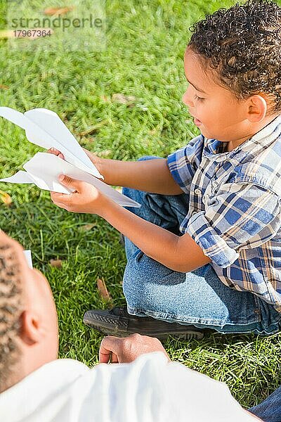 Glücklicher afroamerikanischer Vater und gemischtrassiger Sohn spielen mit Papierflugzeugen im Park