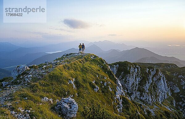 Zwei Wanderer am Gipfel der Benediktenwand bei Sonnenuntergang  hinten Silhouetten von Bergen  Bayerische Voralpen  Bayern  Deutschland  Europa