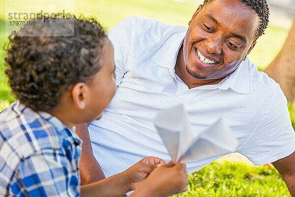 Glücklicher afroamerikanischer Vater und gemischtrassiger Sohn spielen mit Papierflugzeugen im Park