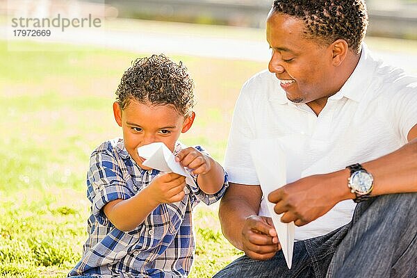 Glücklicher afroamerikanischer Vater und gemischtrassiger Sohn spielen mit Papierflugzeugen im Park