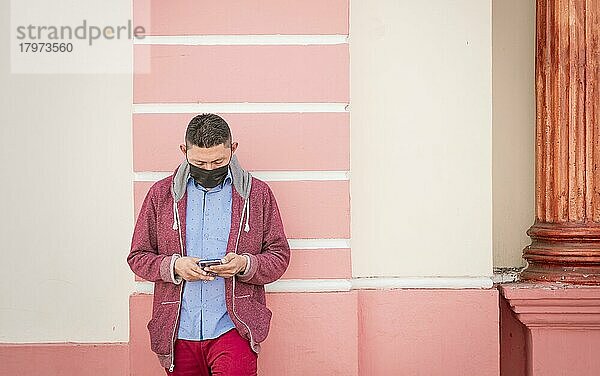 Mann mit Gesichtsmaske  der sein Handy überprüft  junger Mann  der an der Wand lehnt und sein Handy überprüft