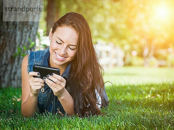 Attraktive  glückliche  gemischtrassige junge Frau  die draußen im Gras liegt und mit ihrem Handy telefoniert