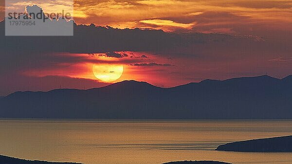 Sonnenuntergang  Sonne groß unter grauen Wolken und direkt über einem Gebirgszug  Landzungen als Schattenriss im Vordergrund  Meer ruhig  Himmel orangerot  Palaiopolis  Insel Andros  Kykladen  Griechenland  Europa